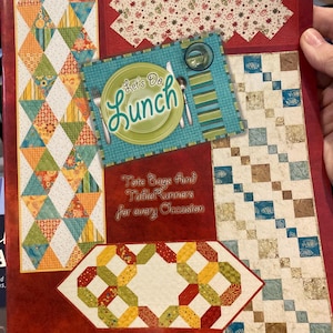 May include: A brochure featuring various quilted table runners and tote bags. The designs include geometric patterns in vibrant colors, a placemat with the text "Let's Do Lunch", and the text "Tote Bags and Table Runners for every Occasion". The Atkinson Designs logo is also visible.