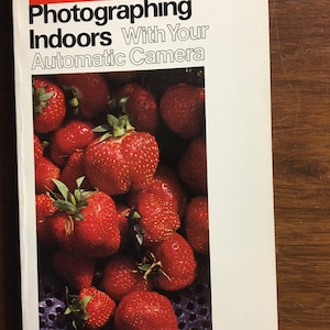 Puede incluir: Una portada de libro con el título "Fotografiar en interiores con su cámara automática" en negrita en negro. La portada presenta una imagen de primer plano de un tazón de fresas rojas.