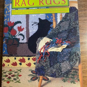 Könnte beinhalten: Buchumschlag mit dem Titel "Rag Rugs" in fetten, schwarzen Buchstaben. Der Umschlag zeigt eine Nahaufnahme eines bunten Teppichs mit einer schwarzen Katzensilhouette und einen Holzstuhl mit einem Teppich aus Stoffresten.