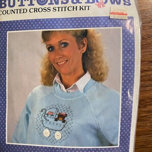 May include: A counted cross stitch kit for sweatshirts, sweaters, or tee shirts. The kit includes a blue canvas, buttons, ribbon, chart, floss, needle, and instructions. The image shows a blue sweatshirt with a cross stitch design of a baby carriage with a teddy bear inside.