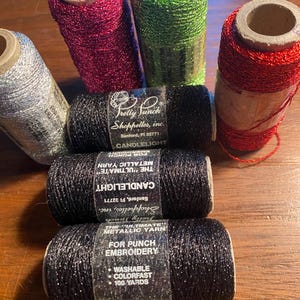 May include: Six spools of metallic yarn for punch needle embroidery. The spools are in various colors, including silver, red, green, black, and pink. Each spool has 100 yards of yarn and is labeled "The Ultimate Metallic Yarn for Punch Embroidery".