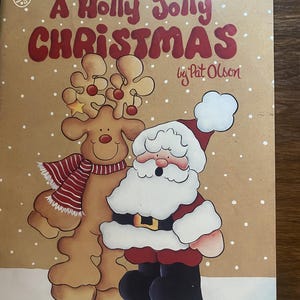 May include: A craft pattern book cover titled A Holly Jolly Christmas by Pat Olson, featuring a tan reindeer with a red and white striped scarf and a cartoon Santa Claus standing in white snow against a tan background with white dots.