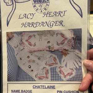 Pode incluir: Um pacote para um projeto Lacy Heart Hardanger. O pacote apresenta um padrão xadrez azul e branco, com imagens de uma placa de identificação, um alfineteiro e uma chatelaine. O texto inclui "Alma Lynne's Works of Art."