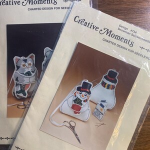 May include: Two cross-stitch kits, one featuring a gray and white cat scissor keeper, the other a snowman scissor keeper. The kits are labeled "Creative Moments" and include charted designs for needlework.