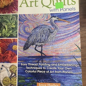 May include: A softcover instructional book titled Creating Art Quilts with Panels by Joyce Hughes. The cover features a large, colorful illustration of a heron standing in water, surrounded by smaller inset images of textured fabric art and quilting.