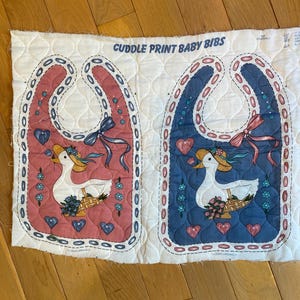 May include: Two baby bibs with a duck design. One bib is pink, the other is blue. Each bib features a white duck wearing a hat, a basket of flowers, and heart accents. The text "CUDDLE PRINT BABY BIBS" is printed at the top.