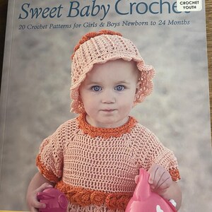 May include: A softcover book titled Sweet Baby Crochet featuring 20 crochet patterns for girls and boys newborn to 24 months. The cover displays a child wearing a peach-colored crocheted bonnet and matching dress with orange trim, holding pink toys.