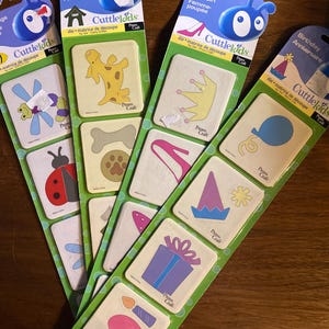 Puede incluir: Varios paquetes de formas troqueladas Cuttlebug, con diseños variados como una jirafa, una corona y una tarta de cumpleaños. Cada paquete tiene un encabezado azul con el logotipo de CuttleKids y texto en francés.