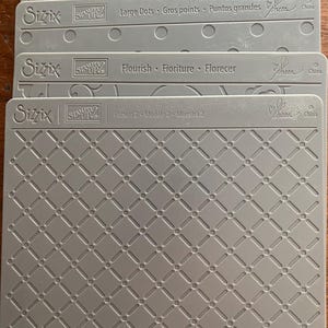 Puede incluir: Una pila de carpetas de relieve Sizzix grises con varios patrones. La carpeta superior está etiquetada como "Harlequin", seguida de "Large Dots", "Flourish" y "Pattern 2". Cada carpeta tiene un diseño diferente, incluyendo triángulos, puntos y una rejilla de diamantes.