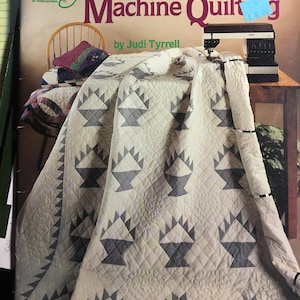 Peut inclure: Un livre intitulé "Guide du débutant pour la couture à la machine" par Judi Tyrrell. La couverture présente une courtepointe avec un motif de panier en noir et blanc sur un fond crème. Le livre est de l'American School of Needlework.