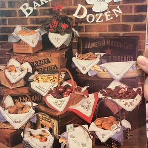 Peut inclure: Une collection de point de croix intitulée "Baker's Dozen" présentant divers paniers et boîtes remplis de produits de boulangerie et de produits. L'image comprend du texte tel que "Ginger" et "James D. Mason Co's". L'arrière-plan est un mur de briques.