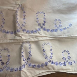 May include: Off-white fabric with blue floral embroidery. The embroidery features a stylized floral design, including a series of small flowers and a larger floral pattern. The fabric appears to be a pillowcase or similar bedding item.