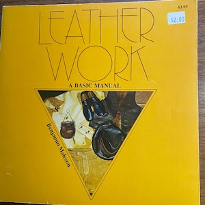以下が含まれることがあります： 「LEATHER WORK」というタイトルの黄色い本で、「A BASIC MANUAL」というサブタイトルが付いています。表紙には革製品の三角形のイラストが描かれています。著者の名前、Benjamin Maleson、が表紙に印刷されています。価格タグには2.00ドルと表示されています。