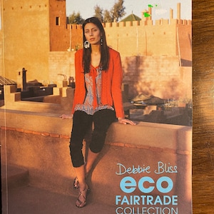 Peut inclure: Une femme portant un cardigan rouge, un pantalon noir et des sandales est assise sur un mur devant un bâtiment. Le texte "Debbie Bliss eco FAIRTRADE COLLECTION" est imprimé sur l'image.
