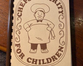 Puede incluir: Un libro de cocina marrón con encuadernación en espiral. La portada presenta un chef de dibujos animados con el texto "Chefs' Charity for Children" en un círculo marrón.