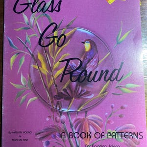 May include: A book titled "Glass Go Round" with a magenta cover. The cover features a painted bird inside a glass circle, surrounded by floral elements. Text includes the title, author names, and "A Book of Patterns".