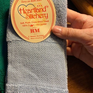 Puede incluir: Una toalla Heartland Stitchery azul claro con una etiqueta. La etiqueta dice "New! Terry on Both Sides" y "Soft, Plush, Cross-Stitch Towel 100% Cotton - 14ct border." La toalla est&aacute; hecha en Hong Kong.