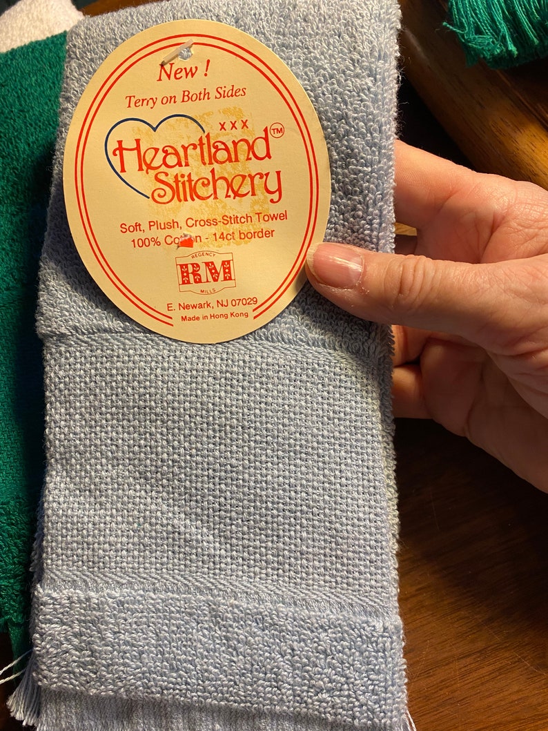 Puede incluir: Una toalla Heartland Stitchery azul claro con una etiqueta. La etiqueta dice "New! Terry on Both Sides" y "Soft, Plush, Cross-Stitch Towel 100% Cotton - 14ct border." La toalla est&aacute; hecha en Hong Kong.