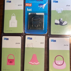 Puede incluir: Seis paquetes de plantillas de troquelado para una máquina de manualidades. Las plantillas son para hacer un delantal, un sombrero, un zapato, un bolso, una falda y un bolso. Los paquetes están etiquetados con el nombre del artículo e incluyen el texto "Quick Kutz" y "Sistema de troquelado personal".