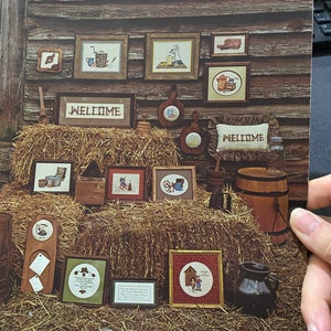 Può includere: Una collezione di schemi di ricamo a punto croce con una varietà di disegni a tema country, tra cui un cartello di benvenuto, un pozzo, una fattoria e un gallo. Gli schemi sono esposti su uno sfondo di legno con balle di fieno e altri decori rustici. Il titolo "The Good Old Days" è stampato nella parte superiore dell'immagine. Il prezzo è di 4,00 €.