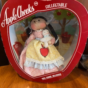 May include: A red apple-shaped box with the words "Apple Cheeks" and "Collectible" printed on it. Inside the box is a doll with a yellow dress, a red apple on her dress, and a white stuffed animal. The doll's name is "Miss Debbie Delicious".