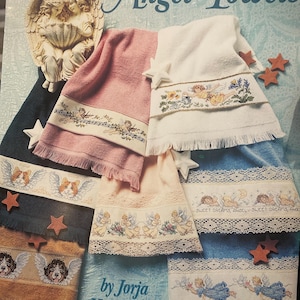 May include: A collection of cross-stitch angel-themed towels in various colors, including pink, white, blue, and tan. Each towel features a detailed embroidered border with angel motifs, floral designs, and the text "SWEET DREAMS, ANGEL". The towels are displayed with star-shaped embellishments.