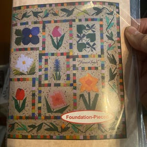May include: A package for a quilt pattern titled "Flower Patch" featuring a colorful floral design. The pattern includes various flower illustrations, with the words "Foundation-Pieced" and the author's name, Marcia L. Baker, printed on the package.