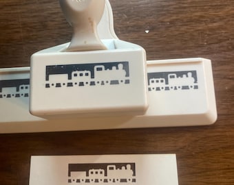 May include: A Martha Stewart craft punch with a train design. The punch is white with a beige handle and creates a train-shaped cutout. A white paper with a train cutout is also visible.