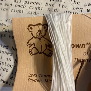 Puede incluir: Primer plano de un hilo de bordar blanco enrollado alrededor de una pequeña tarjeta con una ilustración de un osito de peluche. La tarjeta tiene el texto "3243 Thornv Dryden, Mic" y "own" of Thornv impreso en ella.