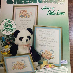 May include: A vintage "Cheesecakes" leaflet with cross-stitch baby announcements. The cover features a panda bear, framed cross-stitch samplers, and wooden blocks. The text reads "Share a Little Love."