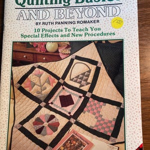 Peut inclure: Un livre intitulé "Quilting Basics and Beyond" de Ruth Panning Romaker. La couverture du livre présente une courtepointe avec une variété de motifs en rose, noir, blanc et marron. La courtepointe est composée de neuf carrés, chacun avec un design différent. La couverture du livre comprend également le texte "10 Projects To Teach You" et "Special Effects and New Procedures".