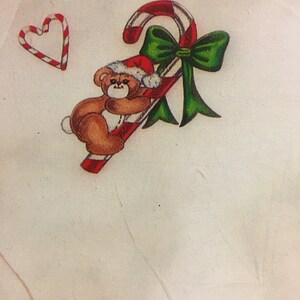 May include: A white sweatshirt with a brown teddy bear holding a red and white candy cane with a green bow. The candy cane is decorated with red and white stripes. There is a red and white candy cane heart in the top left corner of the image.