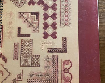 May include: A spiral-bound instructional book titled Jean Hilton's BORDERLINES, featuring a tan cover with various maroon and pink geometric needlepoint border patterns and a solid dark maroon vertical stripe along the right edge of the cover.