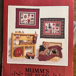 May include: A red and white patchwork quilt sampler with a Christmas theme. The quilt features a variety of traditional Christmas symbols, including a Christmas tree, Santa Claus, a snowman, and a gingerbread man. The quilt is framed in a red and white checked fabric. The quilt is displayed on a shelf with a teddy bear sitting in a wicker basket.