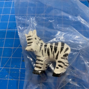 May include: A small, white and black zebra figurine in a clear plastic bag. The zebra is standing on all four legs with its head turned to the side.