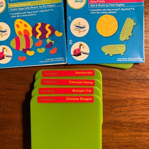 May include: Two packages of Thin Cuts die-cutting templates. The first package is for Easter Eggs and Jelly Beans and the second package is for a ball and boots. There are four green plastic Thin Cuts die-cutting templates stacked on top of each other. The templates are labeled with the names: Hamburger, Carousel Horse, Bumper Car, and Chinese Dragon.
