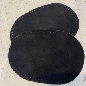 Puede incluir: Dos posavasos ovalados de ante negro con bordes cosidos. Los posavasos están ligeramente desfasados. La costura es de un color más claro que los posavasos. Protegen las superficies del calor y la humedad.