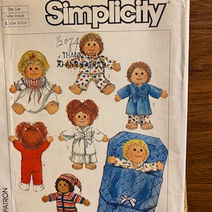 May include: Simplicity sewing pattern featuring doll designs. The pattern includes various doll outfits, such as pajamas, a robe, and a sleeping bag. The dolls have different hairstyles and are in various poses. The pattern is labeled "One Size".