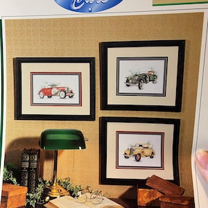 May include: Three framed cross-stitch pictures of classic cars in red, green, and yellow. The frames are black with a cream-colored mat. The top of the image has a blue oval with the words "Classic Cars".