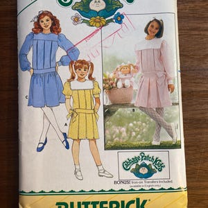 Puede incluir: Patrón de costura Butterick vintage para trajes de Cabbage Patch Kids. El patrón presenta ilustraciones de vestidos en azul, amarillo y rosa. El patrón incluye el texto "FREE PATTERN" y "BONUS! Iron-on Transfers Included."