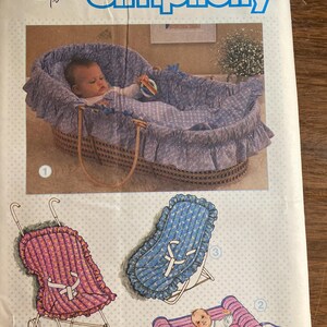 May include: Simplicity sewing pattern featuring a baby in a wicker bassinet with blue floral bedding. Also shown are illustrations of a stroller, a baby seat, and a rolled mat, all with a similar floral pattern.