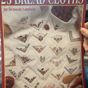 May include: A book cover featuring 25 embroidered bread cloths. The cover has the text "25 BREAD CLOTHS" and "By Deborah Lambein". The cloths are white with various cross-stitch designs, including flowers, hearts, and text.
