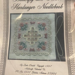 Op de afbeelding: Een naaldenboek met een bloemen- en vlinderontwerp. Het naaldenboek is wit met een kruissteekpatroon van roze bloemen, groene bladeren en vlinders. De tekst "Butterfly and Flower Hardanger Needlebook" staat bovenaan.