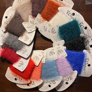 May include: A variety of colors of Wisper embroidery floss, a type of thread used for cross stitch and needlepoint. The floss is arranged in a circle, with each color labeled with a number and letter code.