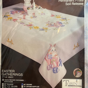 Op de afbeelding: Een witte gestempelde tafelkleed om te borduren, verpakt in plastic. Het ontwerp heeft bloemen- en paashaasmotieven. Inclusief een bijpassende servet met een geborduurde konijn en paaseieren. Verschillende maten zijn beschikbaar.