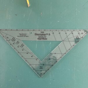 May include: Clear plastic Starmaker 8 quilting tool with measurements and the text "Starmaker 8" and "Kaye Wood". The tool is triangular with angled sides and is designed for quilting. The tool is on a green surface.