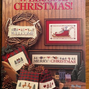 May include: A cross stitch pattern booklet featuring a variety of Christmas-themed designs. The cover features a wreath with a "Welcome" sign, a framed cross stitch of a couple riding in a sleigh, and a cross stitch of a house with the words "Welcome" on it. The booklet is titled "Welcome Christmas!" and is by Peg Wheeler Hope.