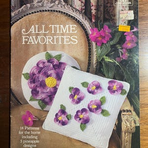 Op de afbeelding: Een vintage boekomslag met de titel "ALL TIME FAVORITES" met haakpatronen. De omslag toont een gehaakte placemat en kussen met paarse bloemmotieven. Het boek bevat 14 huispatronen, waaronder 5 ananasontwerpen.