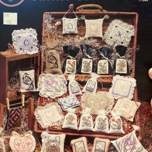 May include: A collection of embroidered sachets and small pillows, featuring floral and initial designs. The image includes the text "Sachet Selections" and "Cross My Heart Inc."