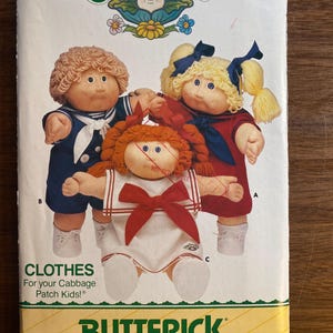 May include: A vintage Butterick sewing pattern featuring Cabbage Patch Kids dolls. The pattern cover shows three dolls in sailor outfits. The text reads "CLOTHES For your Cabbage Patch Kids!" and "BUTTERICK". The background is yellow and cream.